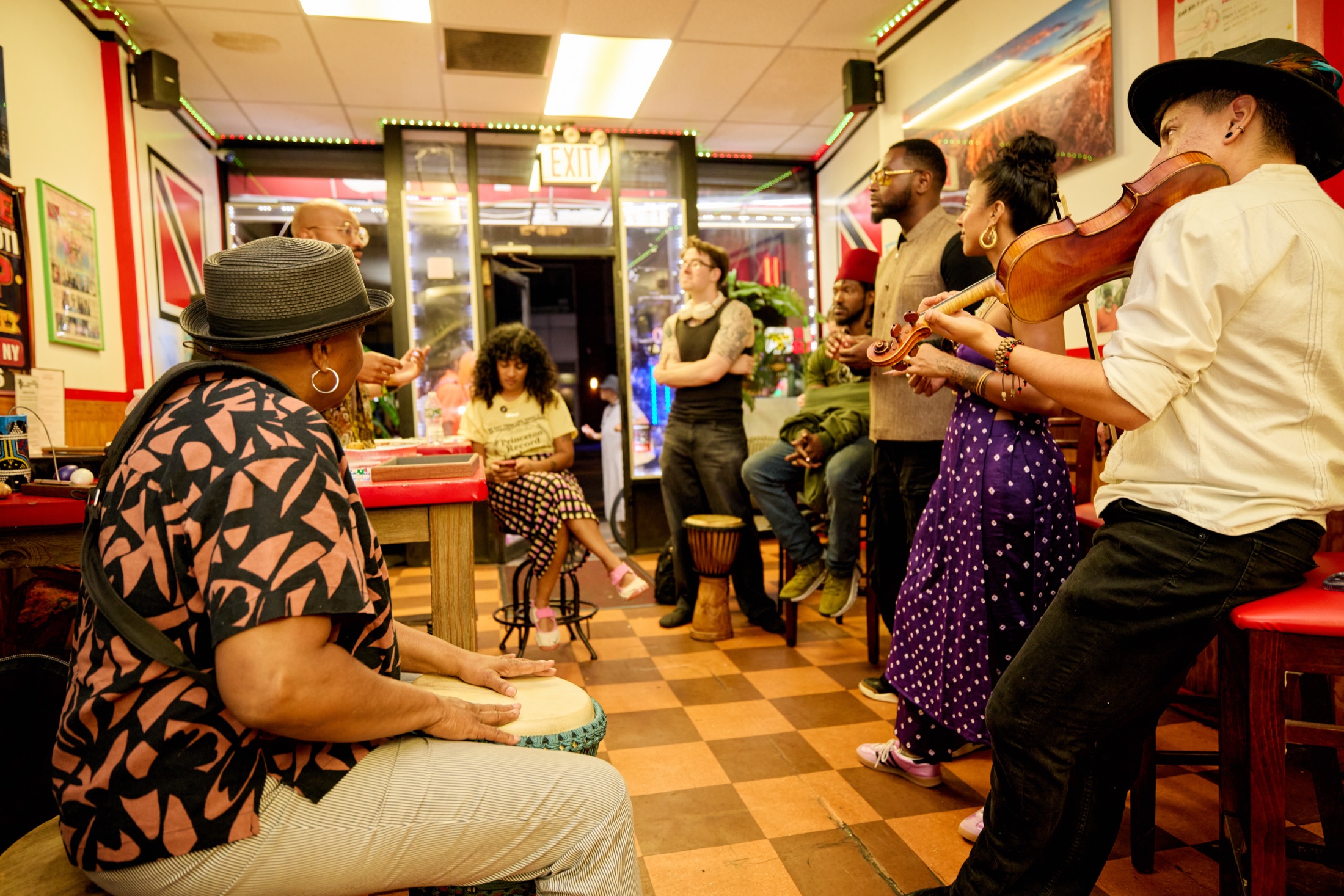 A drummer and a violinist play for a circle of participants standing and sitting around a red table.